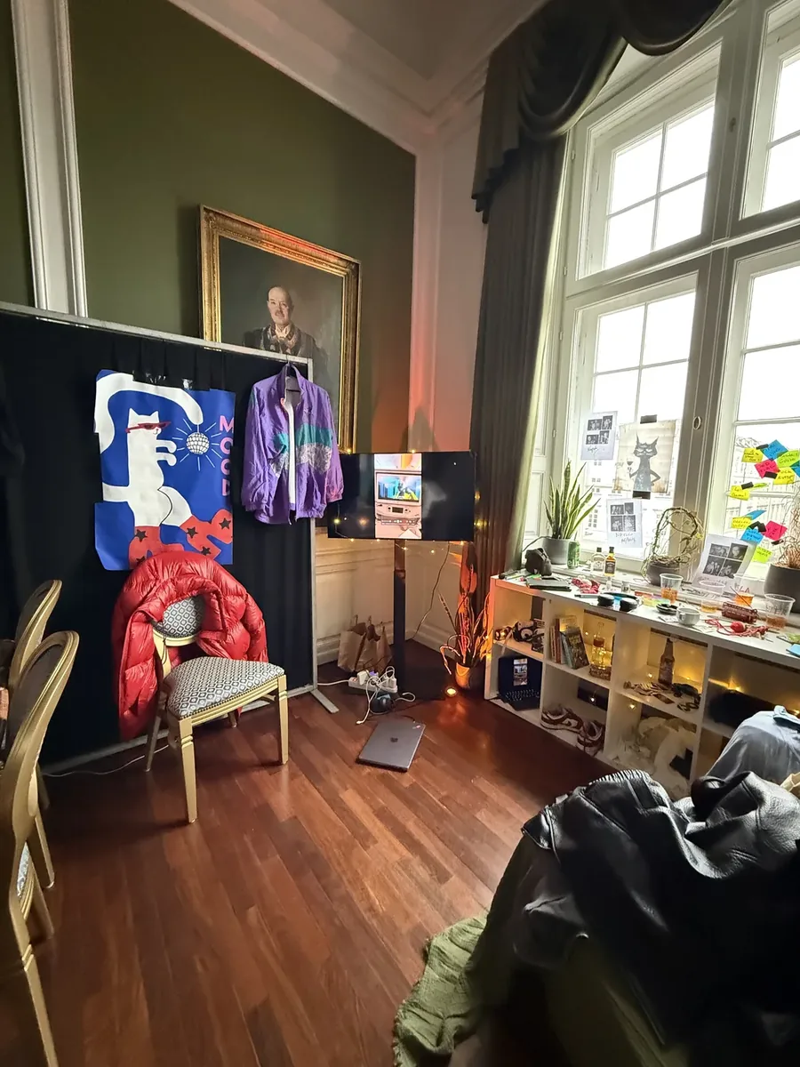 Installation Pandora : écran sur pied dans un salon décoré de vêtements, d'affiches, de plantes et d'objets disposés sur des étagères